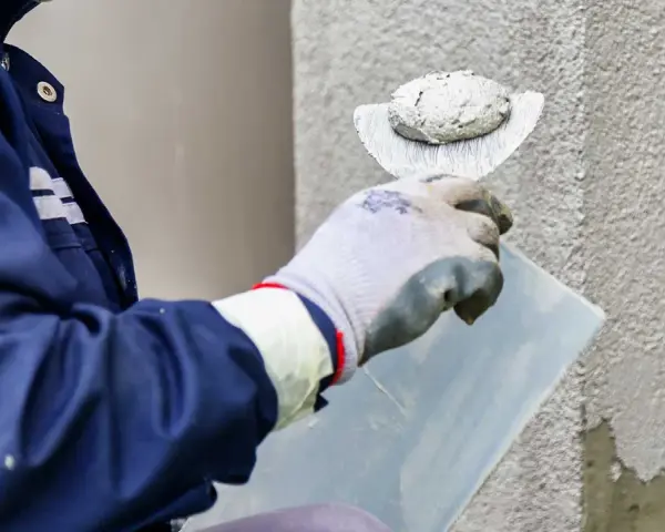 Pracownik w niebieskiej kurtce wykonuje tynkowanie elewacji, nakładając zaprawę kielnią na ścianę budynku.