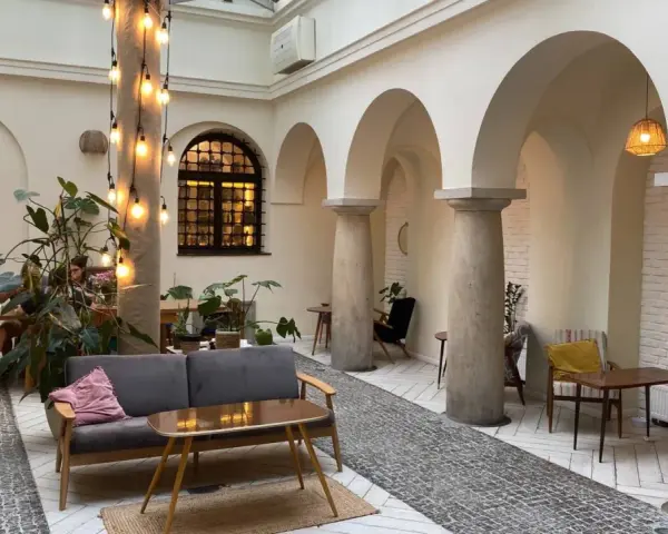 Przytulna kawiarnia patio z łukami i kolumnami. Goście siedzą przy stolikach, a obok stoi wygodna sofa. Widać rośliny i dekoracyjne lampki. Menu czeka na odkrycie.
