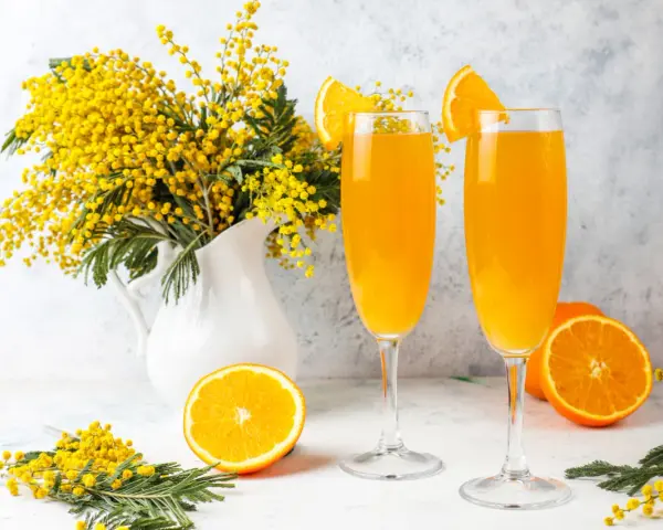 Dwa kieliszki z pomarańczowym napojem, ozdobione plasterkami pomarańczy. Obok bukiet mimozy i połówka pomarańczy. Idealne na wiosenny mimoza drink przepis.