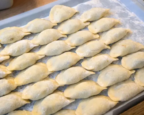 Ułożone na tacy surowe pierogi, posypane mąką.