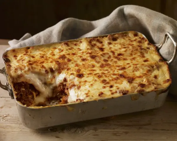 Herzhafte Lasagne mit goldbraun überbackener Käseschicht und einem Hauch von Creme Fraiche, serviert in einer Metallform auf Holztisch.