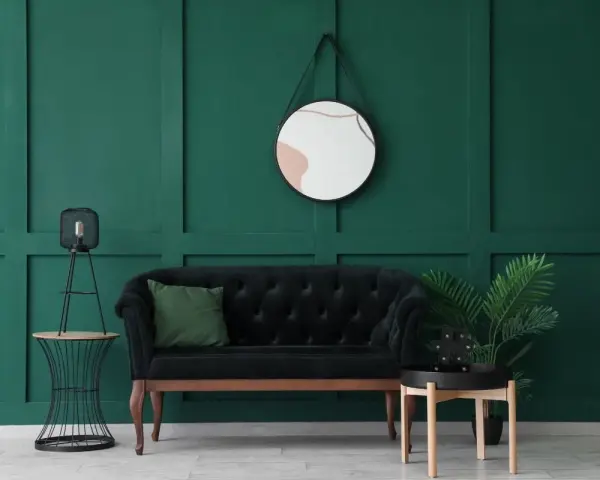 Un elegante divano nero con cuscino verde, un tavolino con lampada e una pianta, e uno specchio dietro il divano su una parete verde scuro.