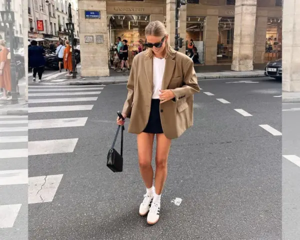 Mujer con look con blazer beige, falda negra y zapatillas blancas cruza un paso de peatones en París.
