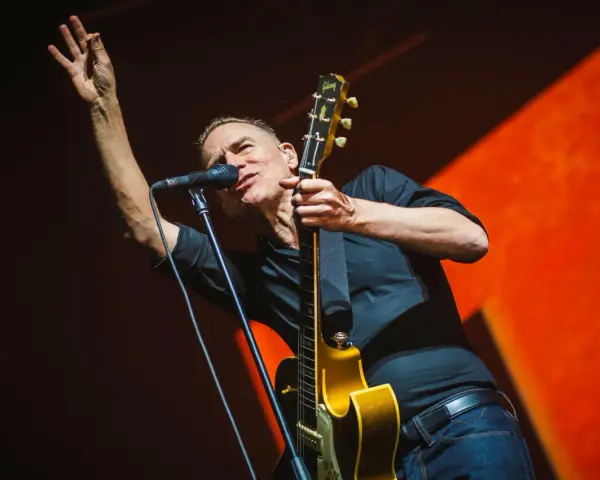 Bryan Adams na koncercie, w czarnej koszuli, z gitarą i mikrofonem, wznosi rękę w geście pozdrowienia.
