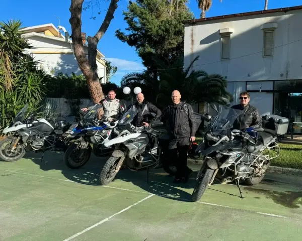 Czterech motocyklistów w pełnym rynsztunku gotowych na **prom na Sycylię**. Motocykle BMW GS zaparkowane na zielonym placu, w tle budynek i palmy.