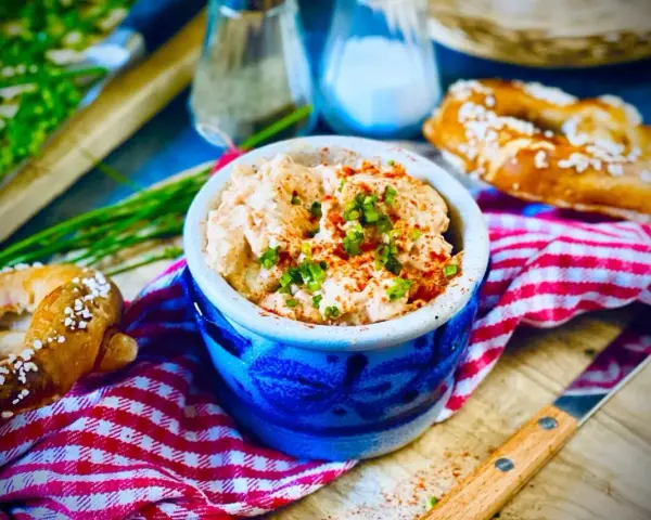 Herzhaftes Obazda-Rezept in einem blauen Steingut-Töpfchen, garniert mit Schnittlauch und Paprikapulver, daneben Brezeln und ein Messer.