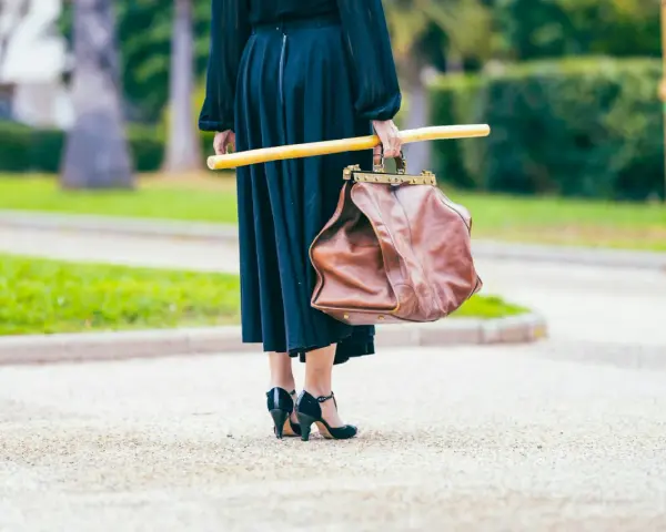 Elegancka kobieta w czarnej sukni i czarnych szpilkach, niosąca brązową torbę i żółty kij. Uosobienie francuskiej mody.