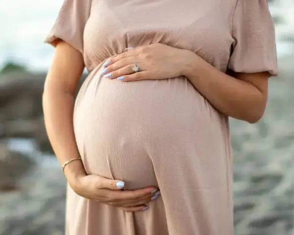 Brzuszek ciężarnej kobiety w beżowej sukience, dłonie delikatnie go obejmują. Warto pamiętać o bezpieczeństwie i unikać stosowania **retinolu w ciąży**.