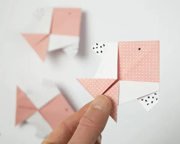 Hand hält Origami-Fisch. Mehrere rosa und weiße Papierfische, die gefaltet wurden, sind im Hintergrund zu sehen.