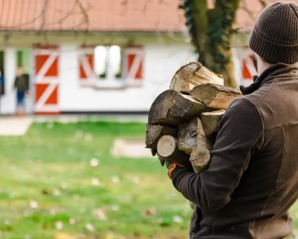 Chcesz wiedzieć, jak zostać leśniczym? Ten mężczyzna z drewnem na podpałkę, w tle dom z czerwonymi okiennicami i dzieci, pokazuje, że praca w lesie to też praktyczne zadania.