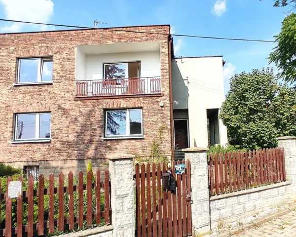 Nowoczesna metamorfoza domu z lat 80. z ceglaną fasadą, balkonem i drewnianym płotem.