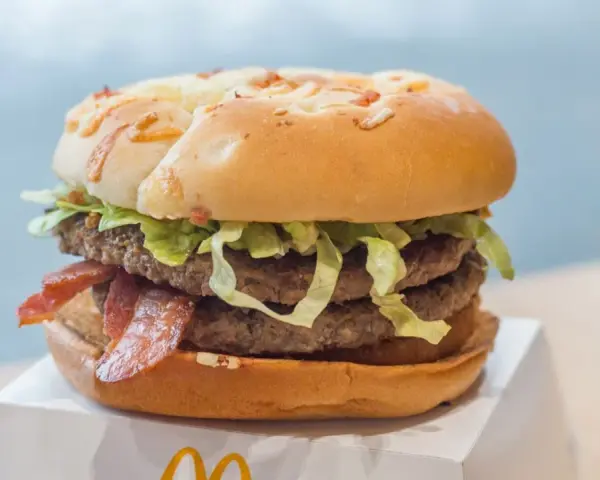 Duży burger z dwoma kotletami, bekonem i sałatą na białym opakowaniu.