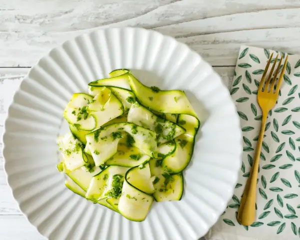 Delicato carpaccio di zucchine condito con erbe fresche, servito su un piatto bianco con forchetta dorata.