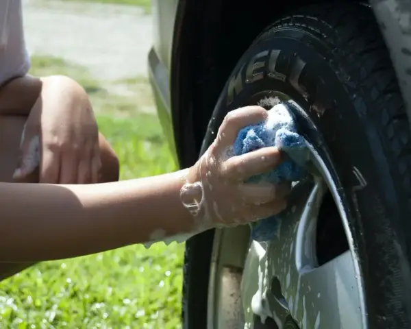 A person's arm and hand, covered in suds, scrub a car tire with a blue sponge. It seems they are testing if you can use dish soap to wash your car.