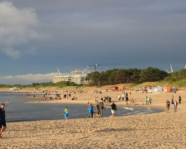 Nadmorskie miasto nad Wieprzą tętni życiem. Ludzie spacerują po plaży, bawią się w wodzie, a w tle widać budowę i wiatraki.