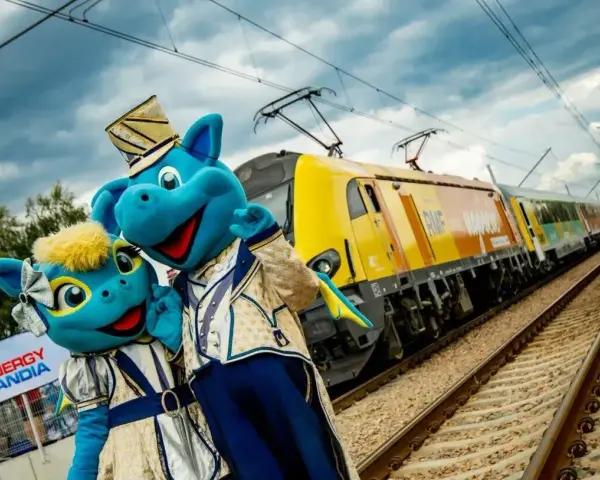 Dwa niebieskie smoki w strojach stoją obok żółtego pociągu na torach kolejowych.