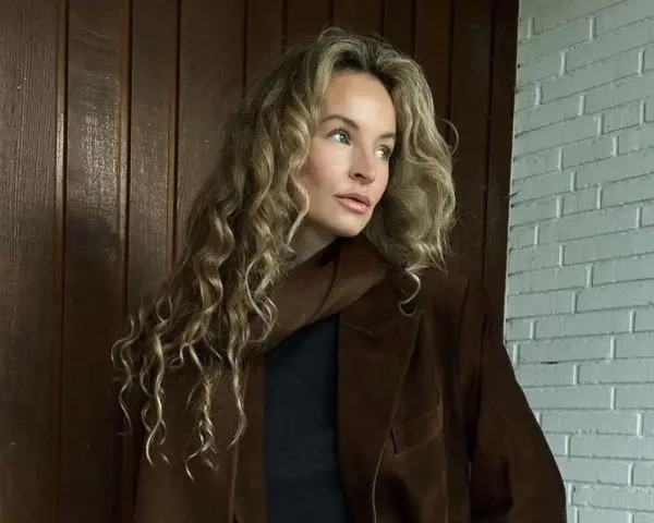 Kobieta z kręconymi, beżowymi włosami, którym pasuje ten odcień. Elegancka stylizacja z brązową marynarką.