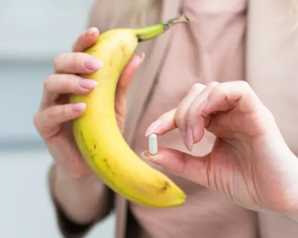 Kobieta trzyma banana i tabletkę, zastanawiając się, ile potasu dziennie powinna przyjmować.