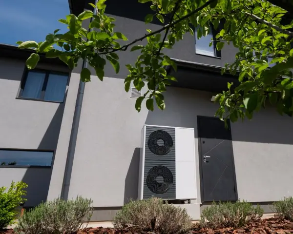 Unità esterna di una pompa di calore, moderna e discreta, installata accanto a una porta su una casa grigia.