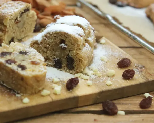 Stollen formen ohne backform: saftige Stollenstücke mit Rosinen und Puderzucker auf Holzbrett, umgeben von Nüssen und Trockenfrüchten.