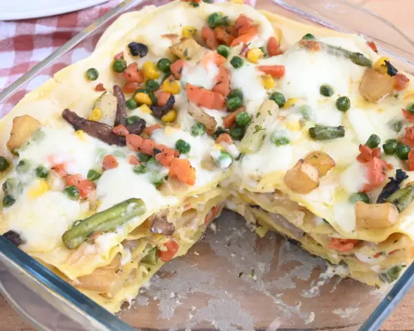 Lasagne di verdure dorate e filanti, con piselli, carote, patate e fagiolini, in una teglia di vetro.
