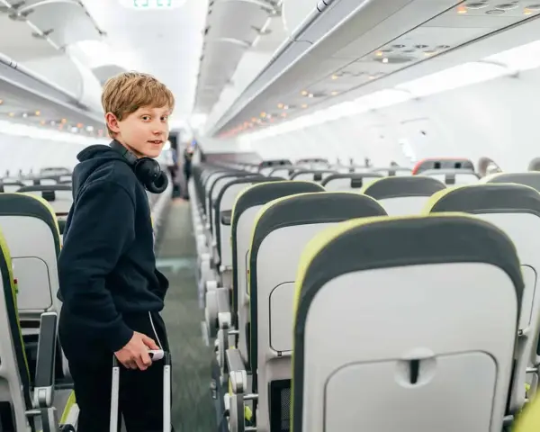Chłopiec z słuchawkami na szyi i walizką w ręku stoi w przejściu samolotu. Od ilu lat można latać samemu? Ryanair pozwala na samodzielne podróże od 12 roku życia.