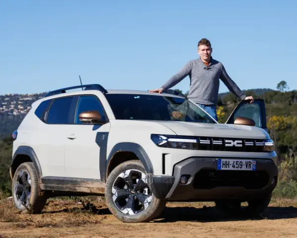Mężczyzna opiera się o maskę jasnoszarego Dacia Duster, hybrydy z fabrycznym LPG, na tle wzgórz.