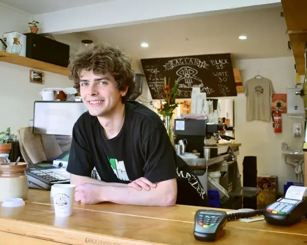 Ein junger Mann mit lockigen Haaren lächelt hinter der Theke eines Cafés. Er scheint seine **zwei Werkstudentenjobs** zu genießen, während er Kaffee zubereitet.