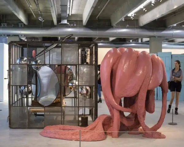 Sculpture rose organique, rappelant l'œuvre de Louise Bourgeois, côtoie une cage métallique remplie de miroirs. Une femme observe l'installation.