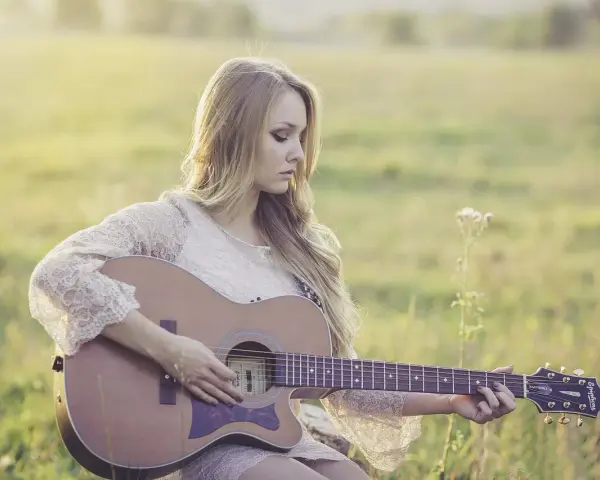 Blondynka w koronkowej sukience gra na gitarze w słonecznym polu. To jak remaster starych wspomnień, pełen ciepła i nostalgii.