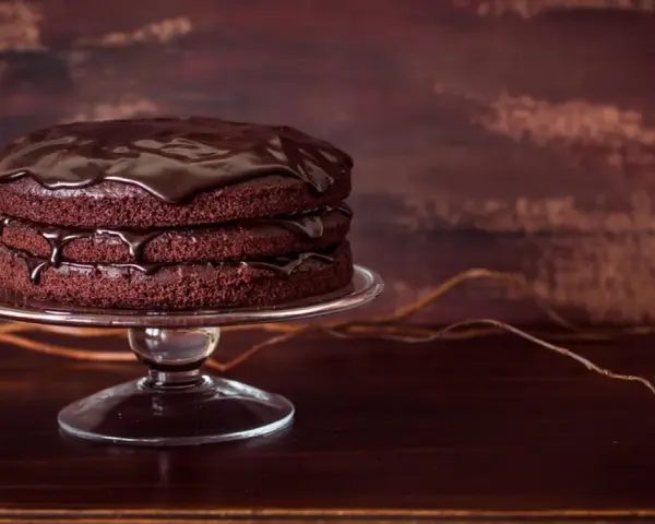 Torta al cioccolato a tre strati, lucida e invitante, su un'alzata di vetro. Gocce di glassa scivolano sui bordi.