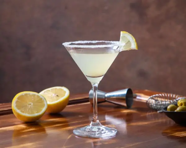 Orzeźwiający drink z martini, udekorowany plasterkiem cytryny i obsypany cukrem. Obok leżą połówki cytryny, oliwki i akcesoria barmańskie. Idealny na domowe drinki z martini.