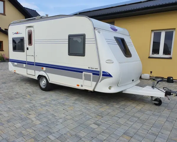 Biała przyczepa kempingowa Hobby 460 UFE "de luxe easy" stoi na kostce brukowej przed żółtym budynkiem.