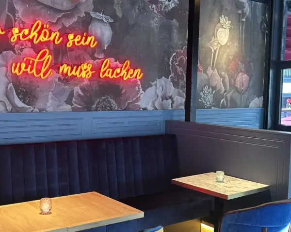 Wilma Wunder: Neonlicht über blauer Bank und floraler Tapete. Wer schön sein will, muss lachen.