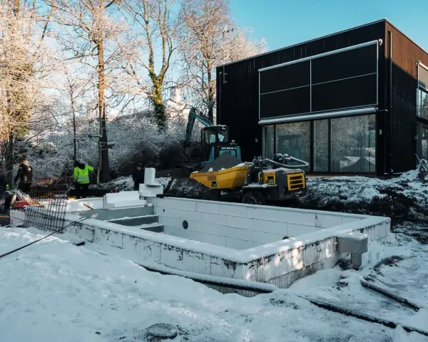 Snö täcker byggarbetsplatsen där en utbyggnad av husets långsida pågår. En grävmaskin och en dumper syns vid en nybyggd poolgrund.