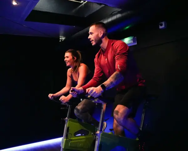 Un homme et une femme s'entraînent sur des vélos d'appartement dans une salle de sport sombre, profitant d'une séance de rpm sport intense.