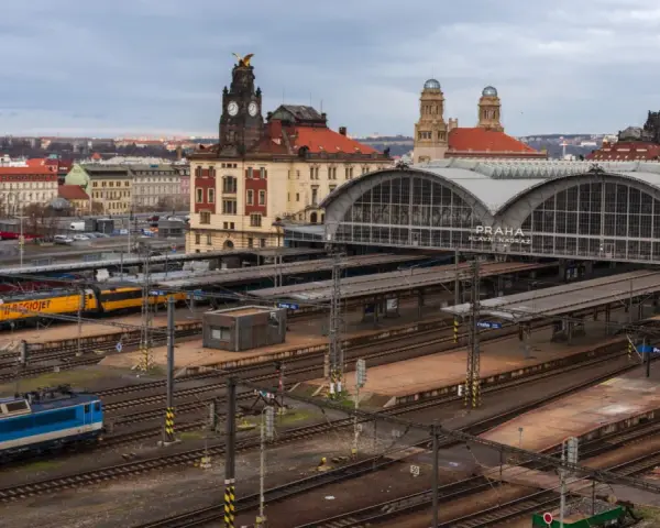 Widok na dworzec kolejowy w Pradze z pociągami na torach i budynkami w tle.