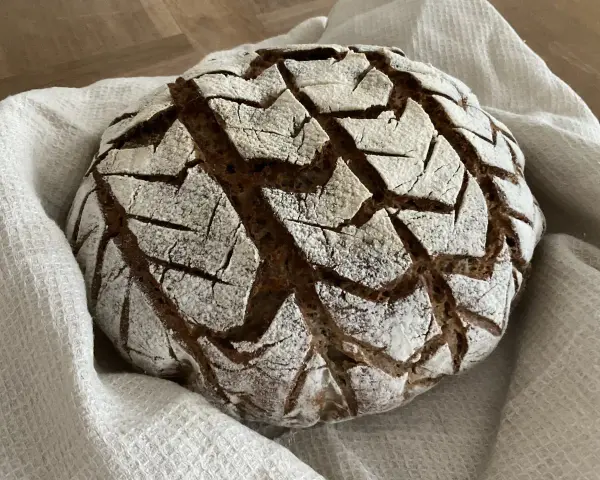 Ein rustikales, rundes buchweizenbrot sauerteig mit einer gemusterten Kruste, bestäubt mit Mehl, liegt auf einem weißen Leinentuch.