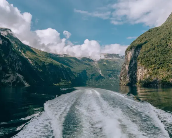 Rejs po Geirangerfjordzie. Pogoda sprzyja, chmury wiszą nad zielonymi zboczami gór, a za statkiem widać biały ślad na spokojnej wodzie.
