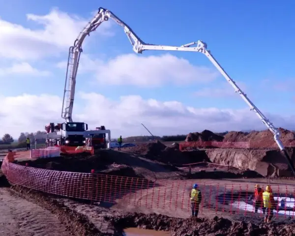 Ogromna pompa do betonu pracuje na budowie, dostarczając materiał do wykopu. Pracownicy w kamizelkach obserwują proces.