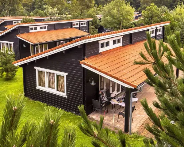 Uroczy bungalow z czarną elewacją i pomarańczowym dachem, otoczony zielenią. Na werandzie stolik i krzesła.