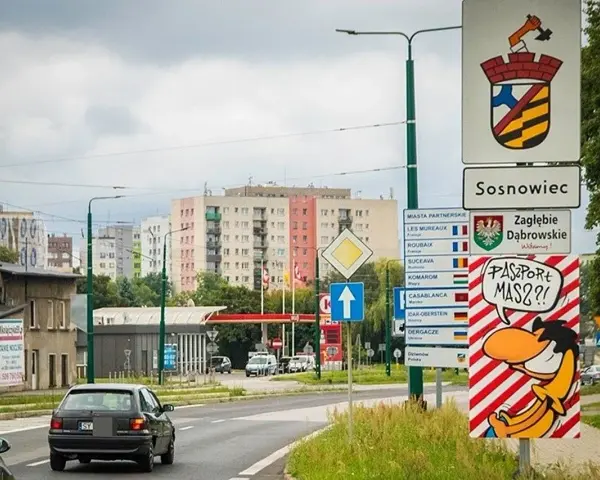 Znak drogowy "Sosnowiec" i humorystyczny napis "Paszport masz?!" przy wjeździe do miasta. W tle bloki mieszkalne.