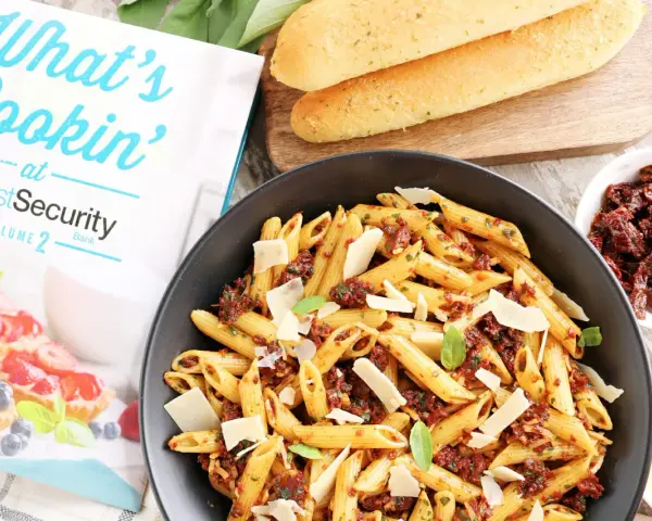 Makaron penne z suszonymi pomidorami, parmezanem i bazylią w czarnej misce, obok pieczywa i książki kucharskiej.