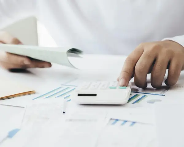 Manos calculando con calculadora blanca sobre gráficos y papeles. Una mano sostiene un talonario, la otra opera la calculadora. Proceso de factura simplificada.