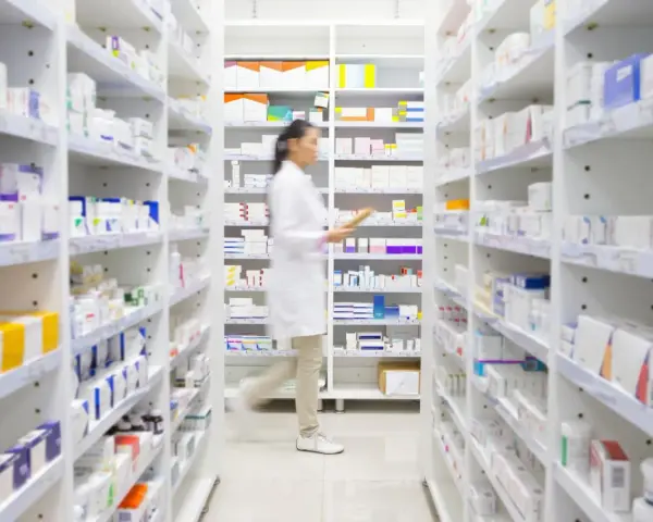 Farmaceutka w białym fartuchu idzie między regałami pełnymi leków, szukając amoksycyliny.
