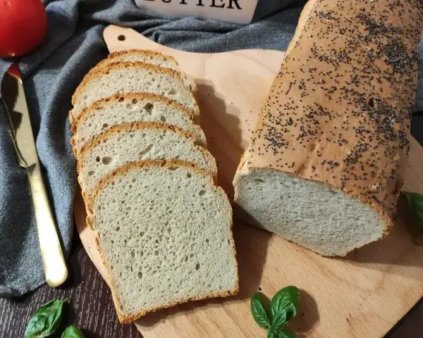 Świeżo upieczony chleb pszenny z makiem, pokrojony w kromki, obok pomidor i złoty nóż.