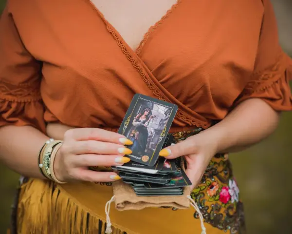 Kobieta w pomarańczowej bluzce trzyma talię kart, na wierzchu widać kartę z cyganką i napisem "3 denary tarot".