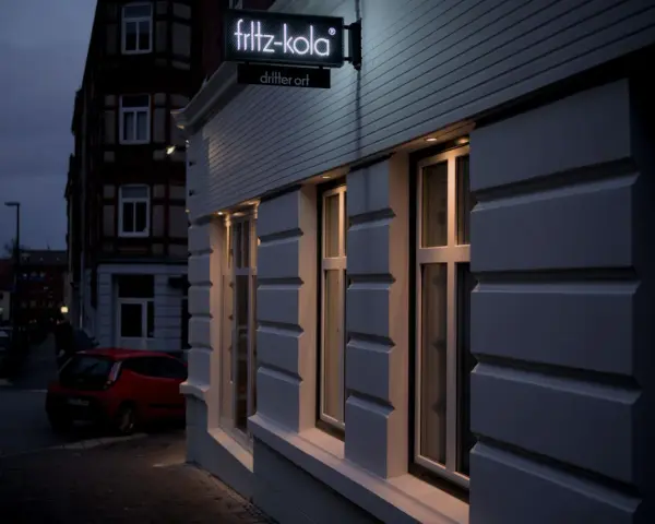 Neon-Schild "fritz-kola" über dem Eingang "dritter ort kiel". Ein rotes Auto parkt auf der Straße.