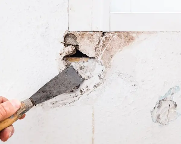 Mano con spatola rimuove intonaco da un buco nel muro troppo grande, rivelando mattoni e detriti.