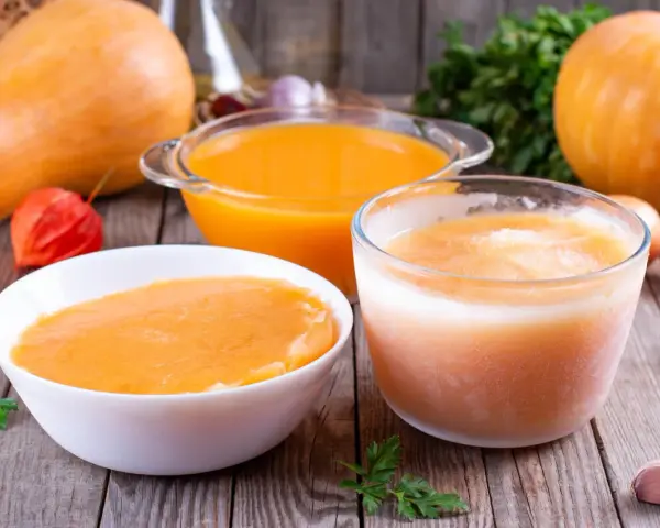 Pomarańczowe zupy w miskach i szklance, dynie i warzywa na drewnianym stole.